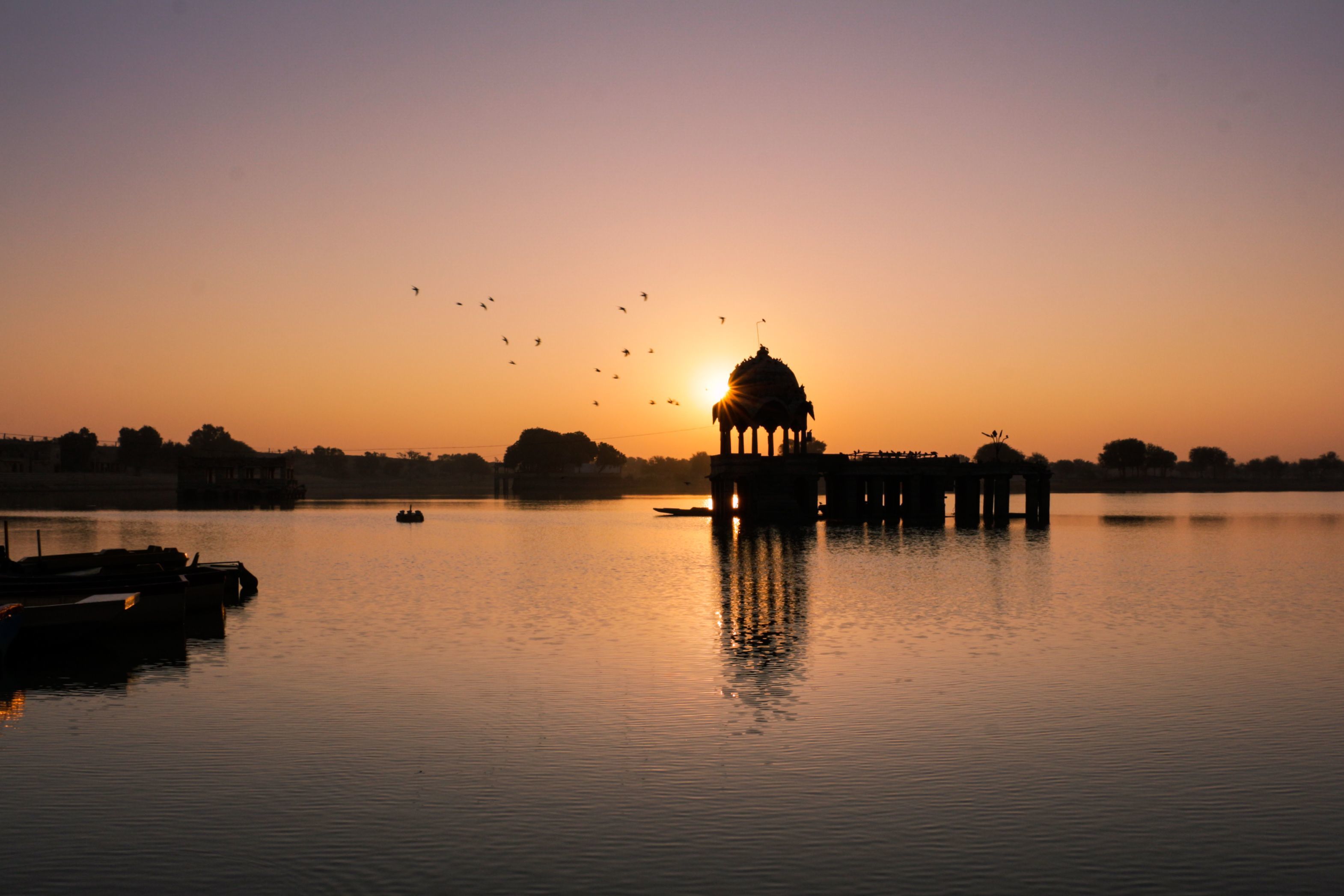 a scenic sunset over a calm gadisar lake featuring a silhouetted temple and flying birds