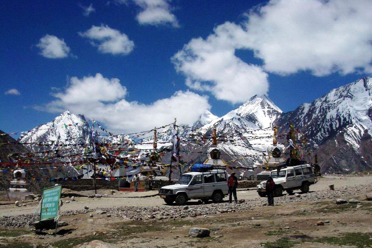 vehicles standing at kunzum pass during daytime with snow capped mountains in the backdrop