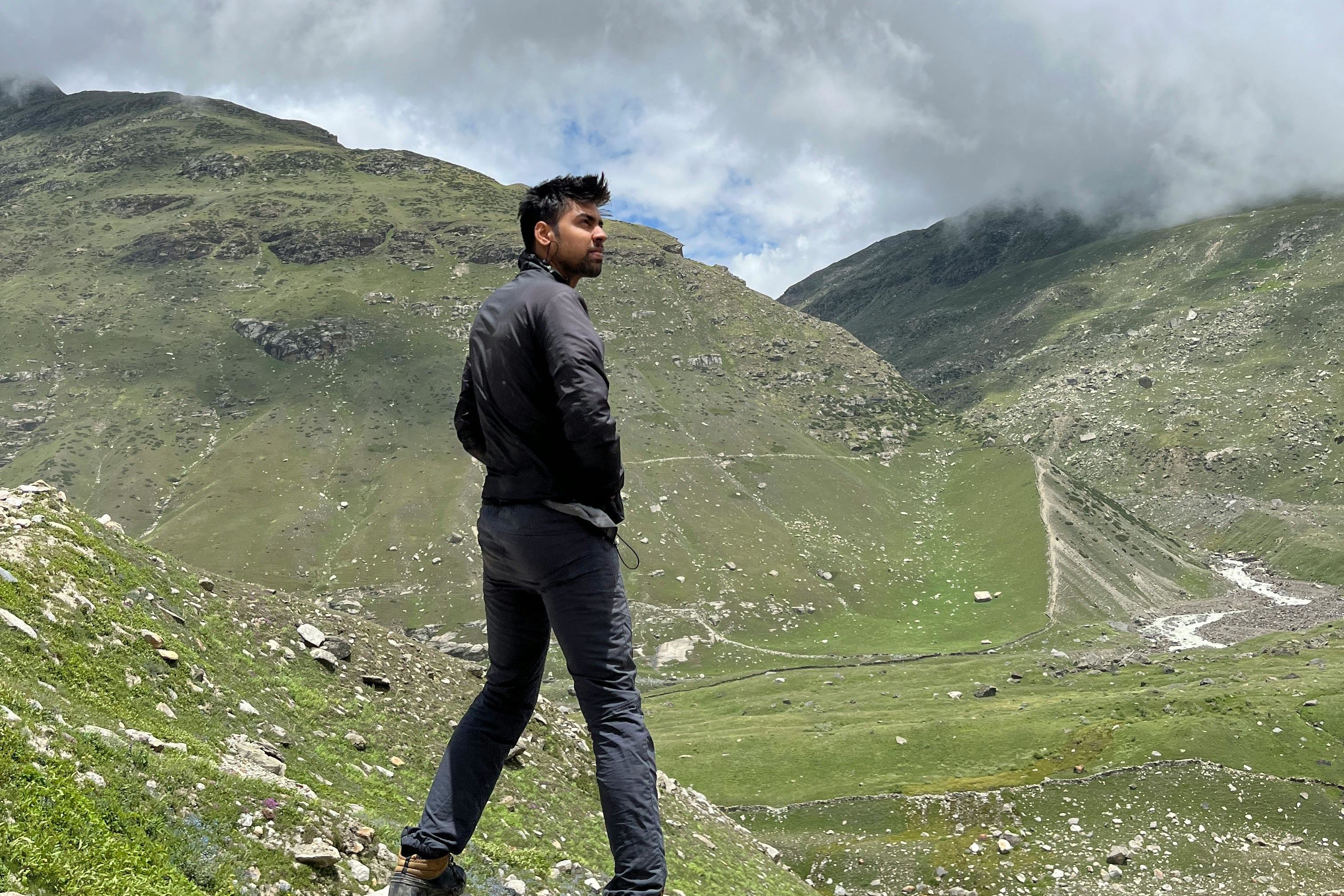 a man in jacket and black pants standing on green grass field under white cloudy sky in lahaul and spiti